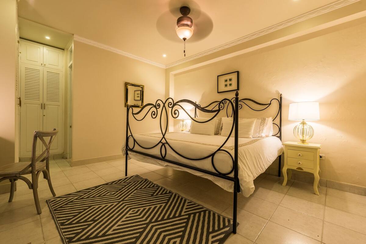 A bedroom with a four-poster bed, a nightstand with a lamp, a chair, and a patterned rug.