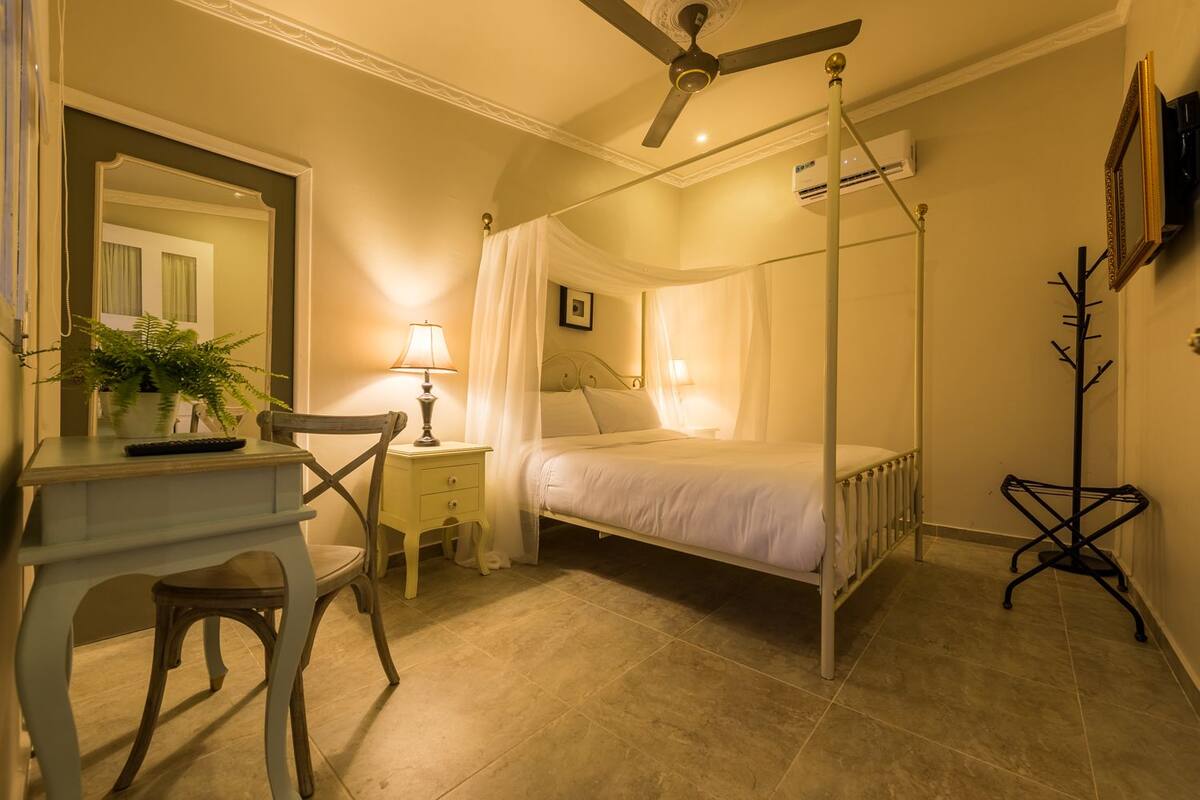 A bedroom with a four-poster bed, a desk with a chair, a bedside table with a lamp, and a coat rack.