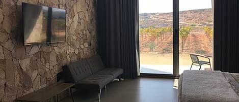 Un dormitorio con pared de piedra, un televisor de pantalla plana, un sofá, una cama y vistas a un viñedo a través de ventanas amplias.