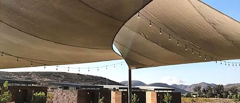 Un área de comedor al aire libre con mesas y sillas bajo grandes toldos, con vistas a un paisaje montañoso.