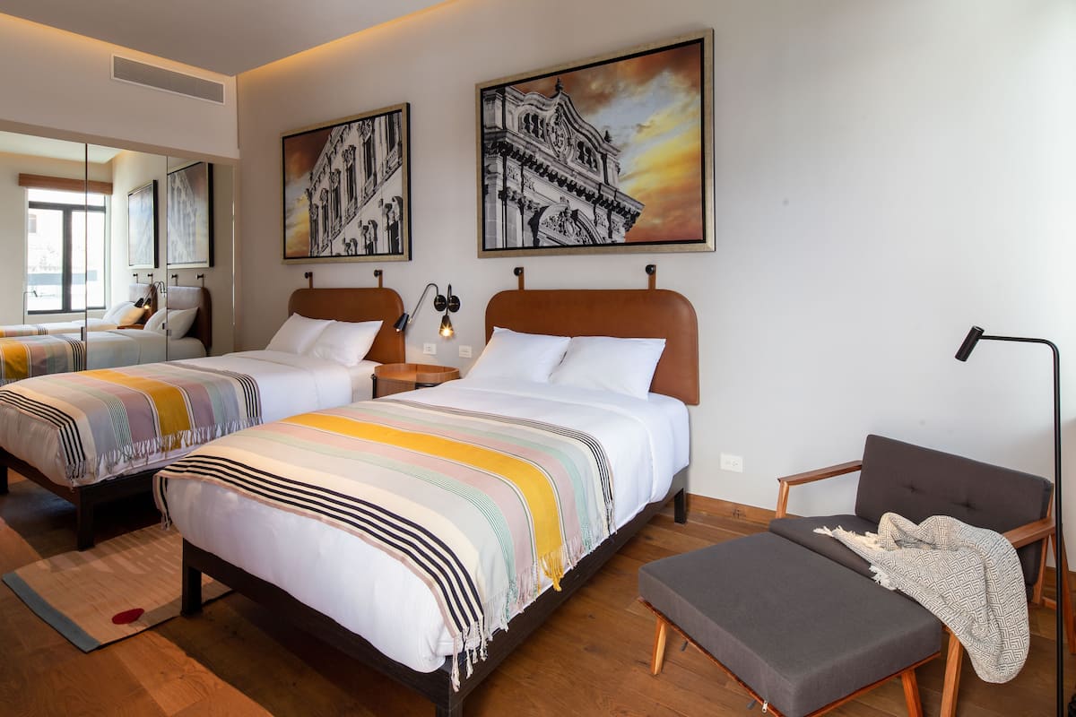Camera d'albergo con due letti, un divano, una scrivania e una sedia. Due quadri alle pareti e una finestra con vista.