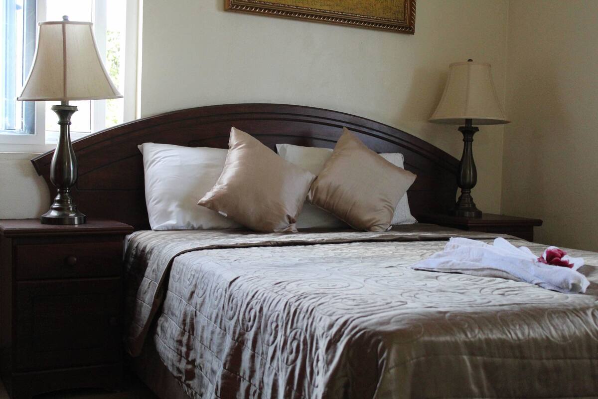 A neatly made bed with a brown headboard, two bedside lamps, and a window with curtains.