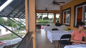 Un espacio comedor al aire libre techado, con mesas y sillas, un ventilador de techo y vistas a la calle y zonas verdes.