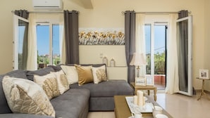 A living room with a grey sofa, decorative pillows, a coffee table with cups and a framed picture, and a view through large windows. - Four seasons House (Corfu)