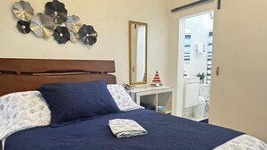 Un dormitorio con cama, un espejo en la pared, una mesita con un objeto rojo y blanco, y una puerta que da al baño.