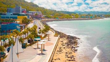 A coastal promenade with a sandy beach, a rocky shoreline, and a row of buildings along the water's edge.