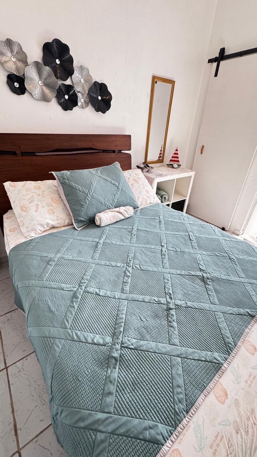 Uma cama com colcha acolchoada, cabeceira de madeira e mesa de cabeceira com espelho e pequeno objeto decorativo.
