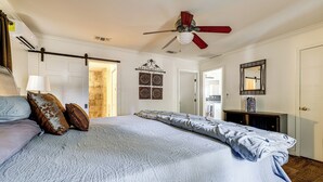 Een slaapkamer met een groot bed, een plafondventilator, een spiegel aan de muur en een deur naar een andere kamer.
