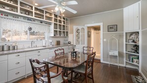 A kitchen with white cabinets, a dining table, and chairs. There is a ceiling fan, a window with lace curtains, and a shelf with decorative items. - Charming Victorian Farmhouse, Privacy, Wi-FI , 5 min to Durango (Durango)