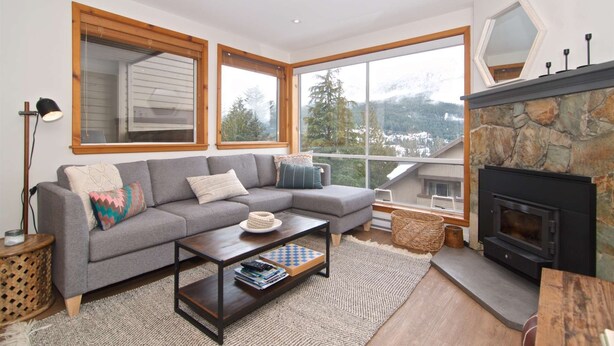 Una sala de estar acogedora con un sofá seccional gris, una mesa de centro de madera y una chimenea de piedra. Amplias ventanas ofrecen vistas a un paisaje montañoso nevado.