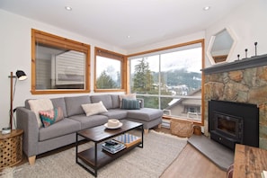 A cozy living room with a grey sectional sofa, a wooden coffee table, and a stone fireplace. Large windows offer a view of a snowy mountain landscape. - PRIVATE Hot Tub spacious family townhouse. 3 Levels, Carport, Creekside location (Whistler)