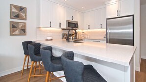 Una cocina moderna con armarios blancos, un refrigerador de acero inoxidable, un microondas y una cafetera. Hay tres taburetes de bar con cojines oscuros.