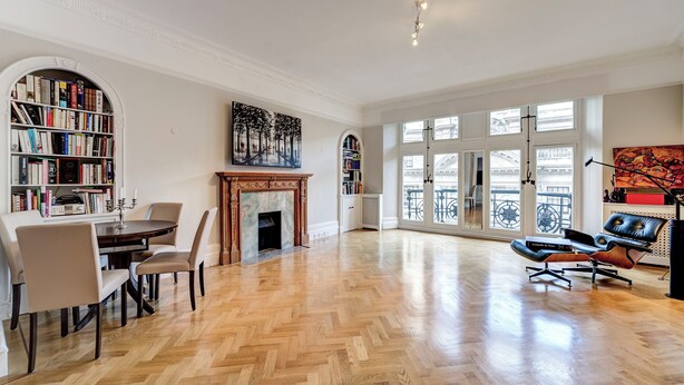 Une pièce spacieuse dotée d’un parquet en bois disposé en chevrons, d’une cheminée, d’une bibliothèque, d’un coin repas avec des chaises et d’une grande fenêtre offrant une vue dégagée.