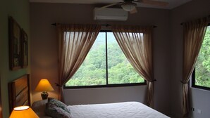 Ein Schlafzimmer mit Bett, Nachttisch, Lampe, zwei Fenstern mit Vorhängen und einem Deckenventilator.