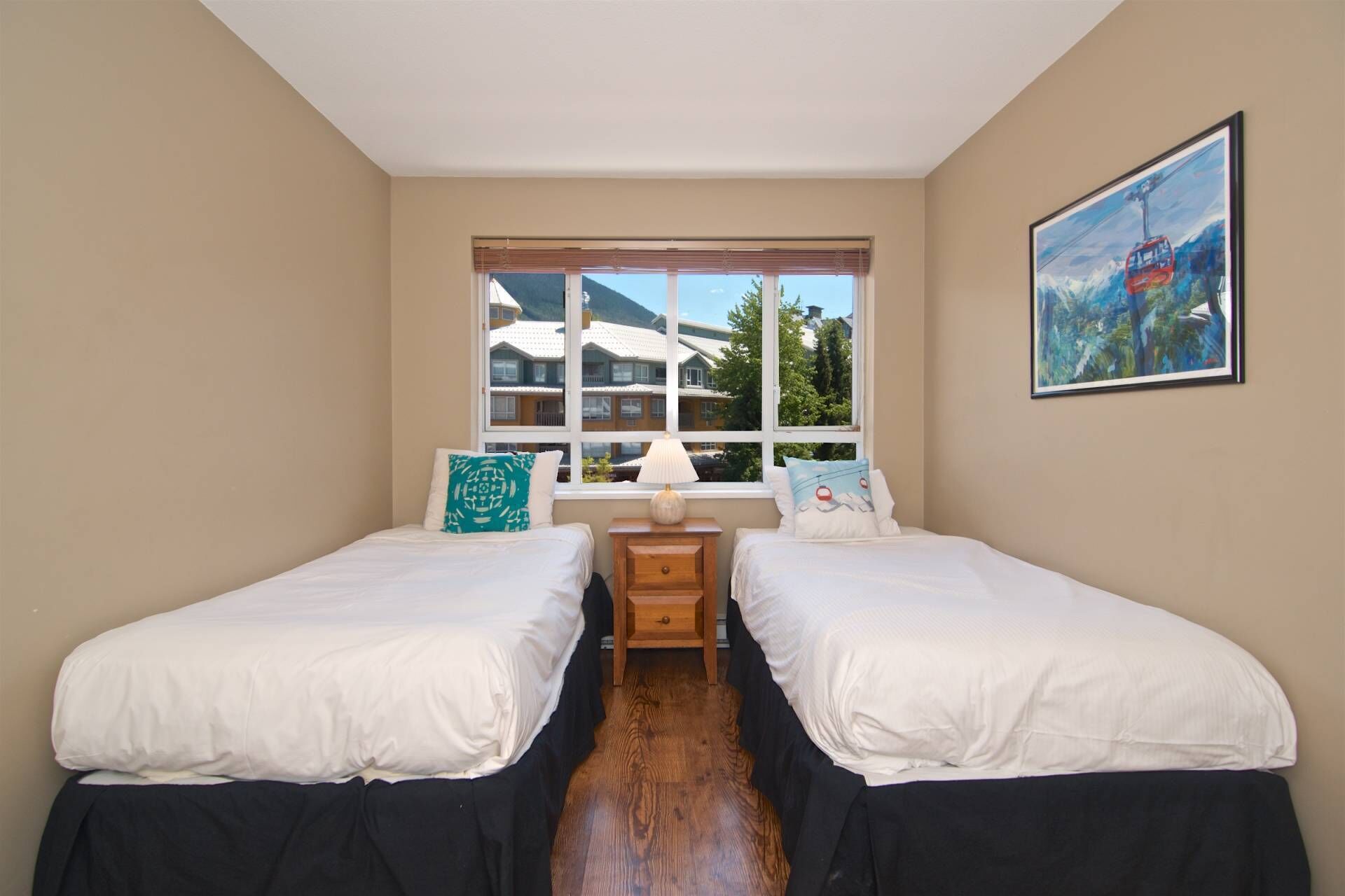 A room with two beds, a window with a view of buildings, a small table with a lamp, and a painting on the wall.