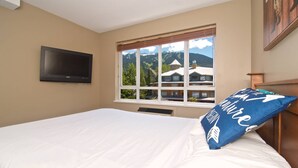Ein Schlafzimmer mit Bett, einem an der Wand befestigten Fernseher und einem Fenster mit Blick auf Häuser und Berge.