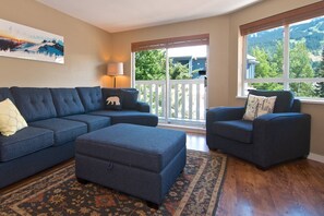 A living room with a sectional sofa, a loveseat, and a matching ottoman. There is a large window with a view of a deck and trees.