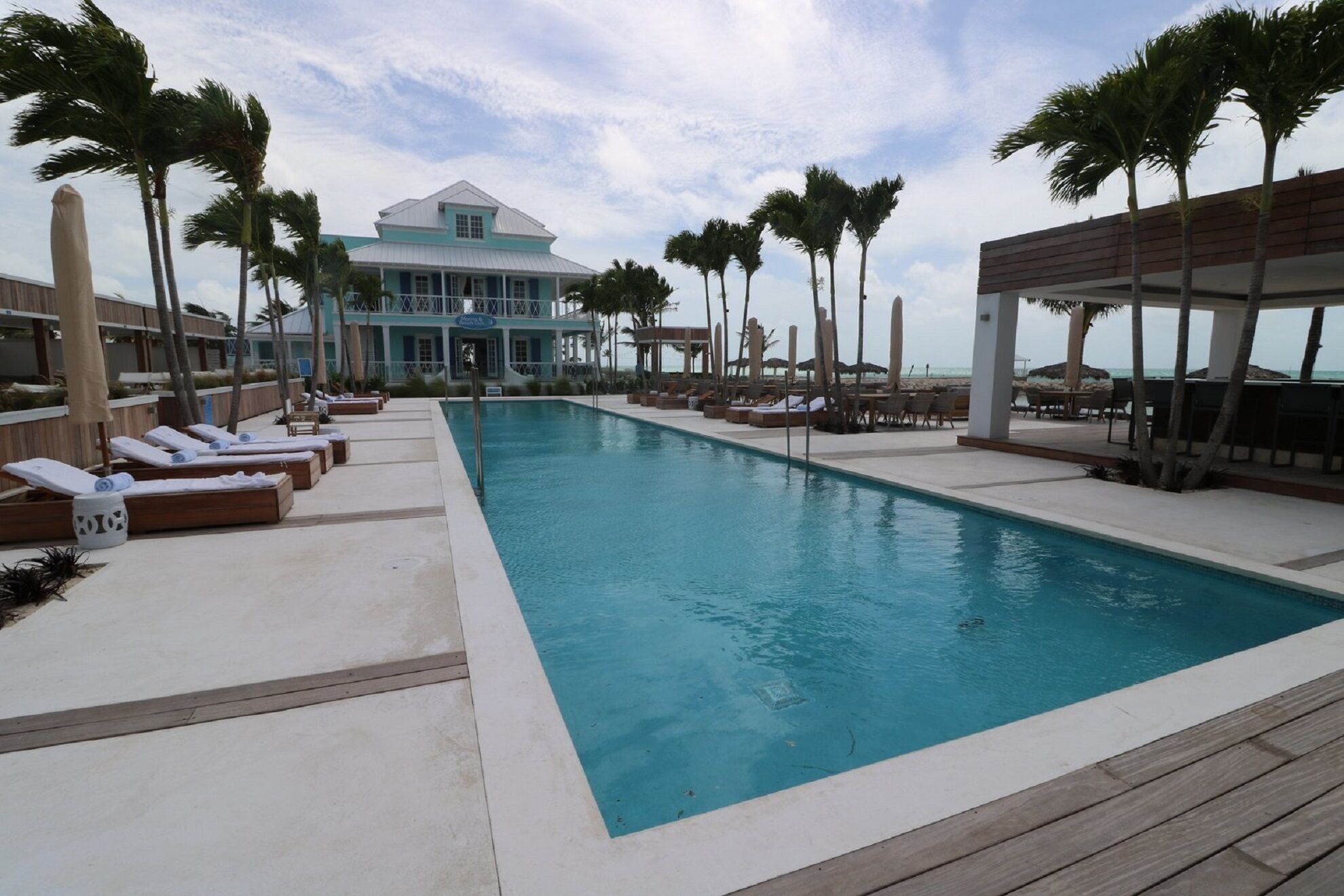 A poolside area with lounge chairs and umbrellas, a building with a blue roof, and palm trees.