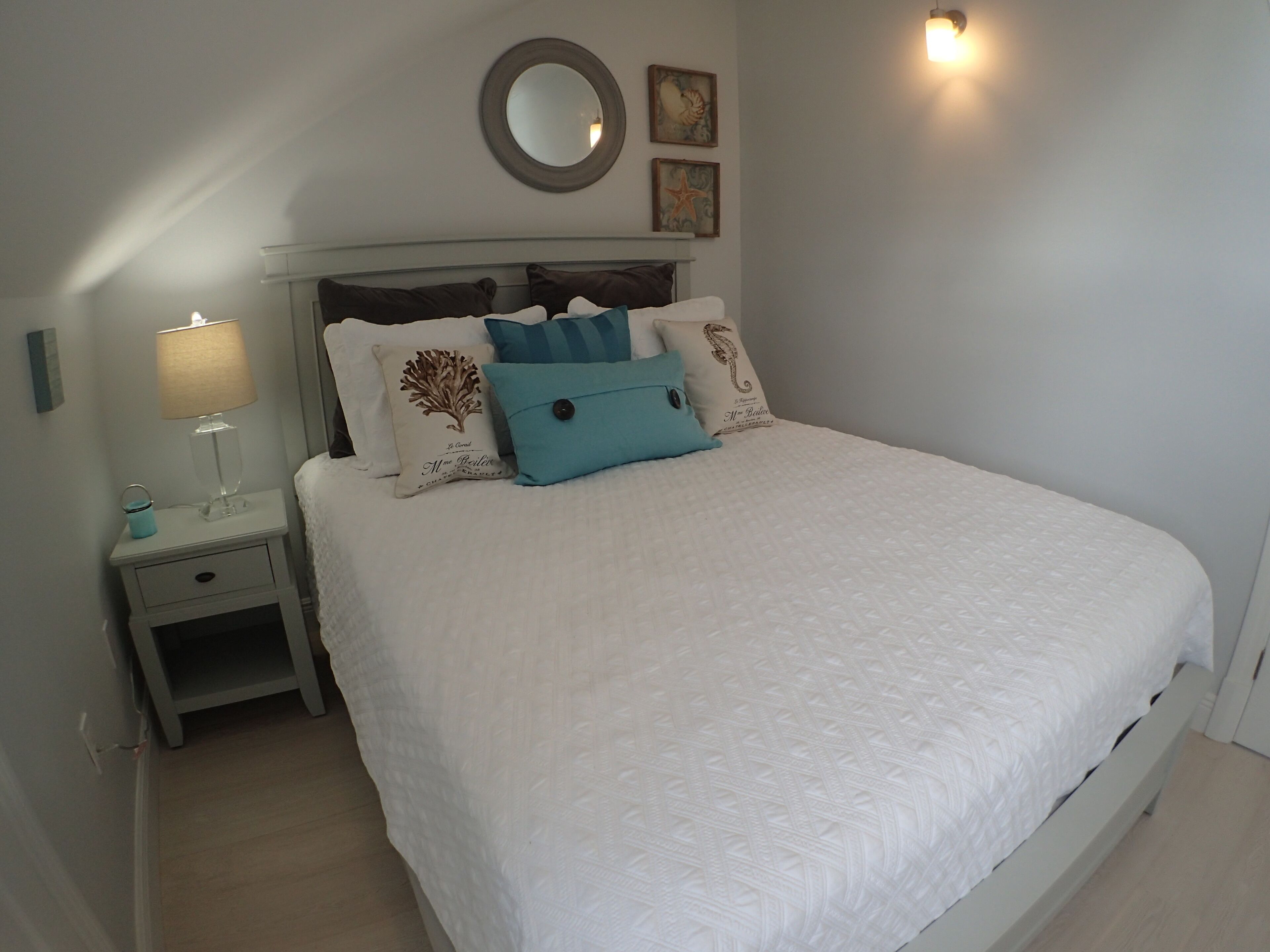 Un lit bien fait, recouvert d’une couette blanche, une table de chevet avec une lampe et un miroir fixé au mur.
