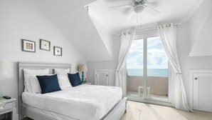 Un dormitorio con cama, mesita de noche, ventilador de techo, ventana con cortinas y vista al mar.