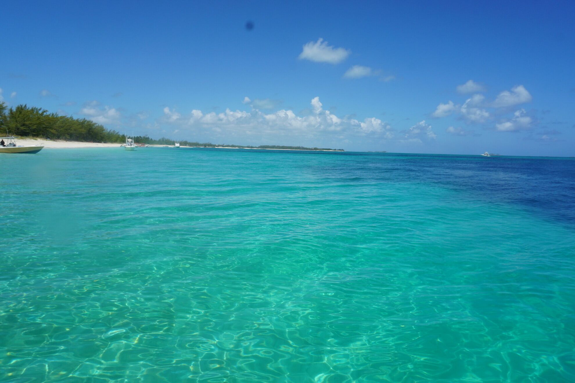 A clear turquoise sea with a boat on the left and a beach with trees on the left side.