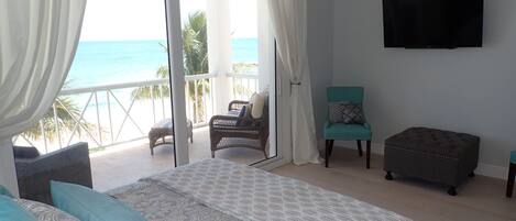 Quarto com cama, cadeira, banco, televisão e varanda com vista para o mar.