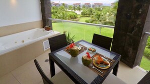 Un balcón con una mesa de vidrio dispuesta para comer, una bañera y vistas a un paisaje verde con edificios al fondo.