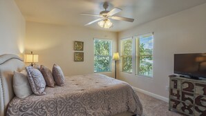 Ein Schlafzimmer mit einem Bett, Deckenventilator, Fernseher, Kommode und zwei Fenstern mit Jalousien.