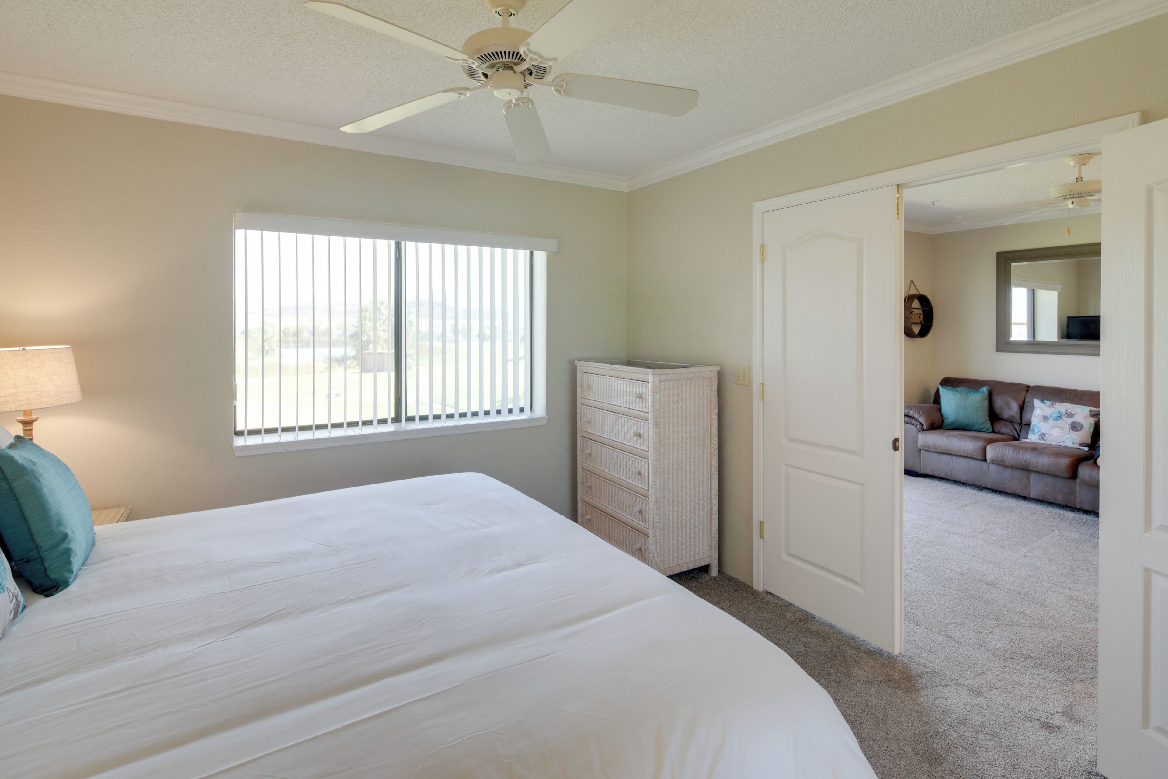 Un dormitorio con cama, cómoda, ventana con persianas, ventilador de techo y puerta que da a otra habitación.