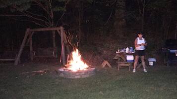 Une personne se tient près d’un feu de camp dans une zone boisée, à proximité d’une balançoire en bois et d’une table sur laquelle se trouvent divers objets.
