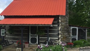 Une cabane rustique en rondins de bois, avec un toit rouge et des murs en pierre, dotée d’un porche avec des meubles d’extérieur et des parterres de fleurs.