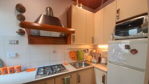 Een keuken met een gasfornuis, oven, magnetron en koelkast. Er zijn kastjes, een spoelbak en diverse keukenbenodigdheden.