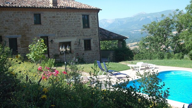 A stone house with a swimming pool, surrounded by lush greenery and mountains in the background.