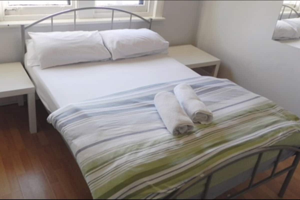 Ein Zimmer mit einem Einzelbett, Metallgestell, weißer Bettwäsche und einer gestreiften Decke. Es gibt einen kleinen Nachttisch und ein Fenster über dem Bett.