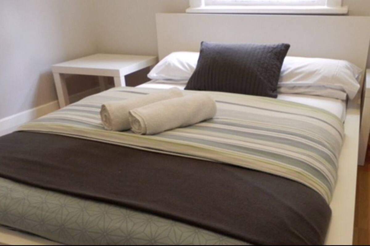 Ein ordentlich bezogenes Bett mit gestreifter Bettdecke und gerolltem Handtuch, zwei Nachttische und ein Fenster über dem Bett.