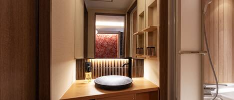 Un baño moderno con un tocador de madera, un lavabo redondo y un espejo encima. Se ve una zona de ducha a la derecha.