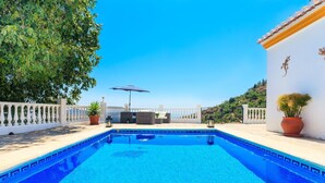 Uma piscina azul límpida cercada por um muro branco, plantas em vasos e vista para uma colina.