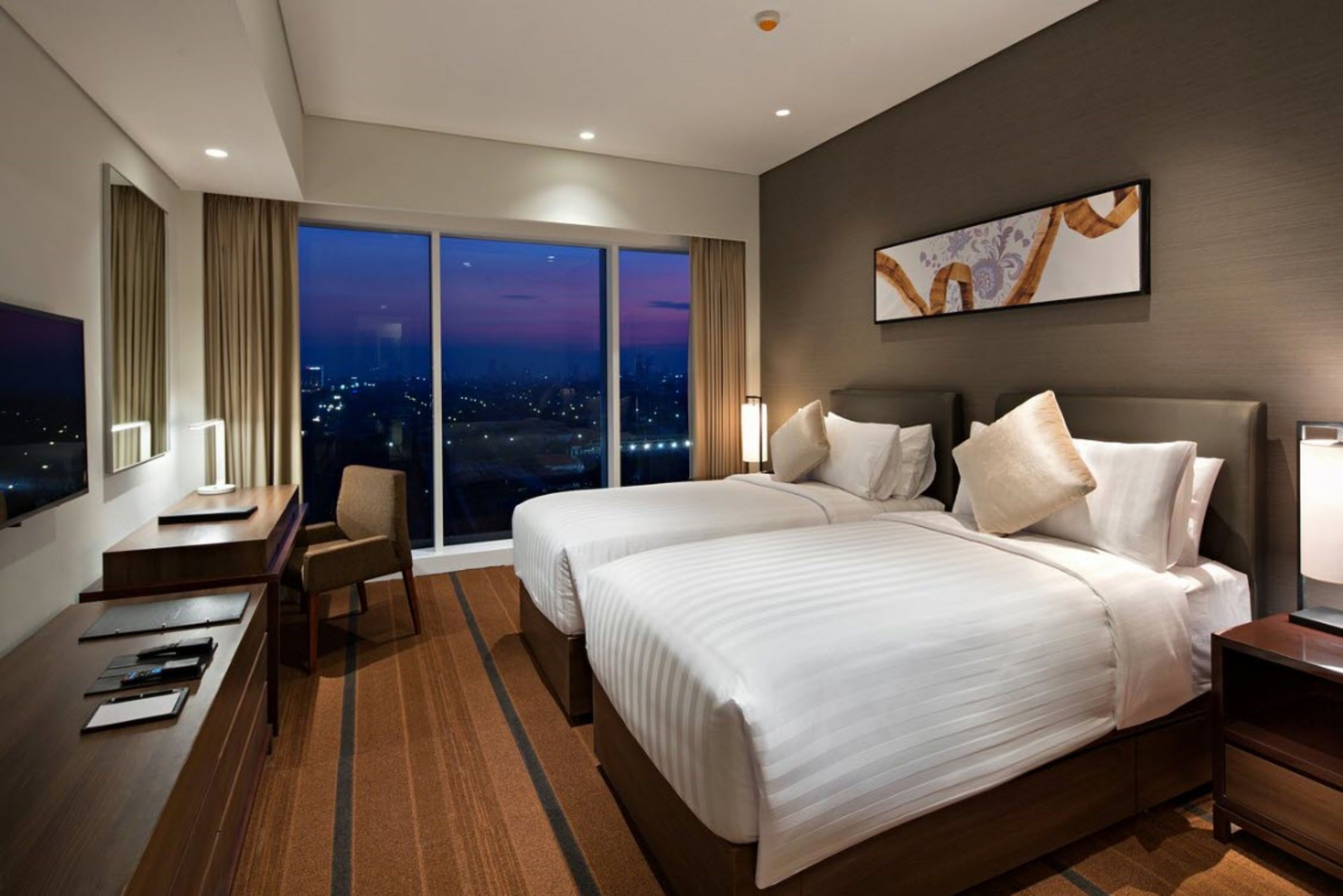 A hotel room with two beds, a desk, a chair, and a large window with a city view.