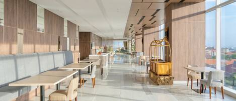 A modern dining area with long tables and chairs, a central seating area, and large windows offering a city view.
