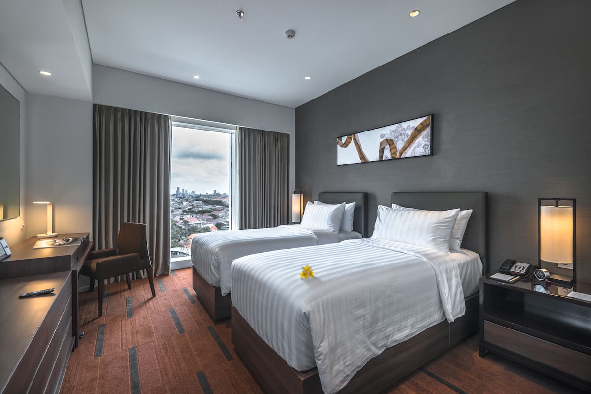 A hotel room with two beds, a desk, a chair, and a large window with a city view.