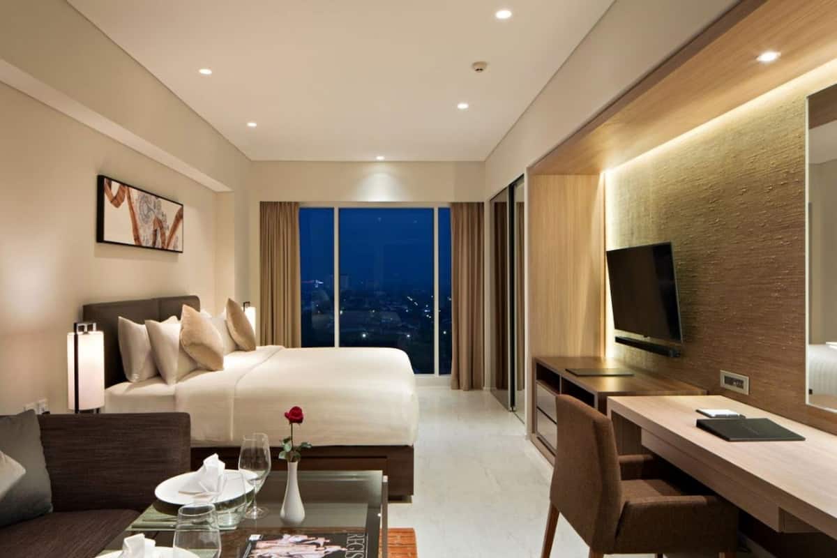 A hotel room with a bed, a desk, a chair, a dining table, and a large window with a city view.