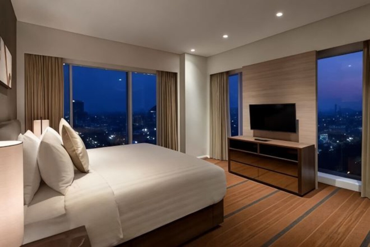 A modern hotel room with a large bed, a TV on a wooden stand, and a city view through large windows.