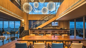 A modern interior with a large wooden table, comfortable seating, a bar area, and a staircase leading to an upper level.