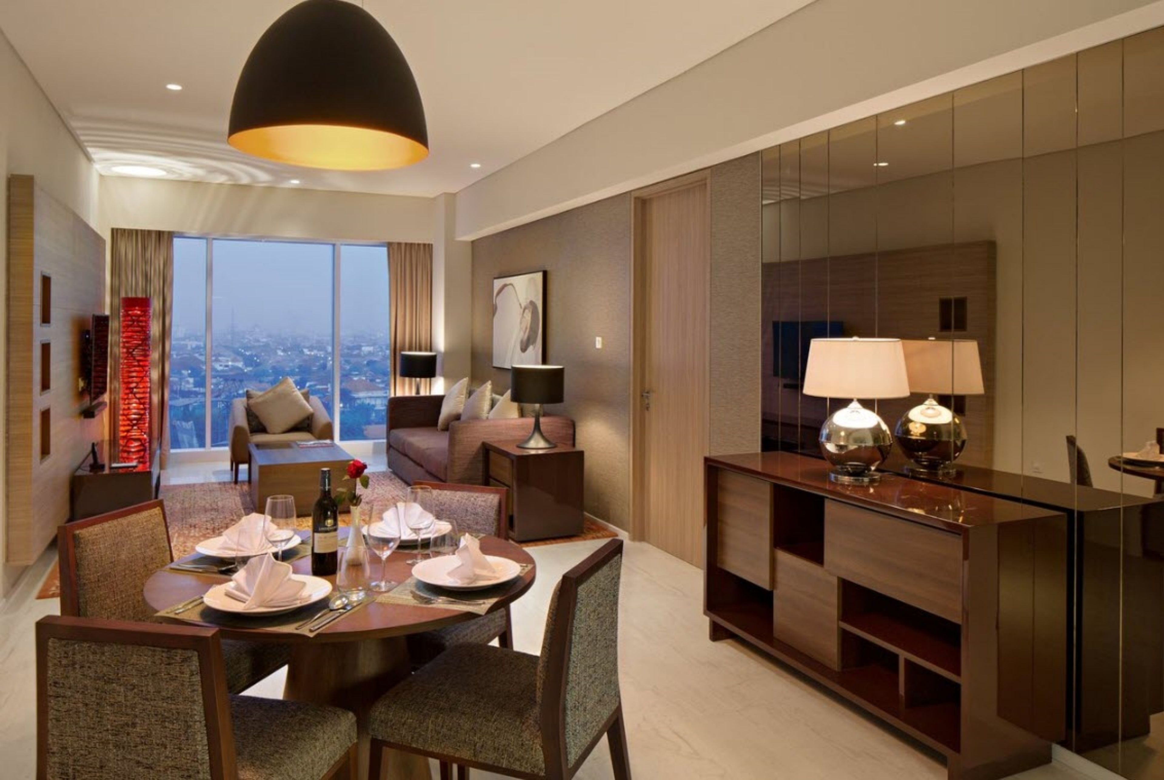 Quarto de hotel moderno com área de refeições, sofá, espelho grande e vista para a cidade.