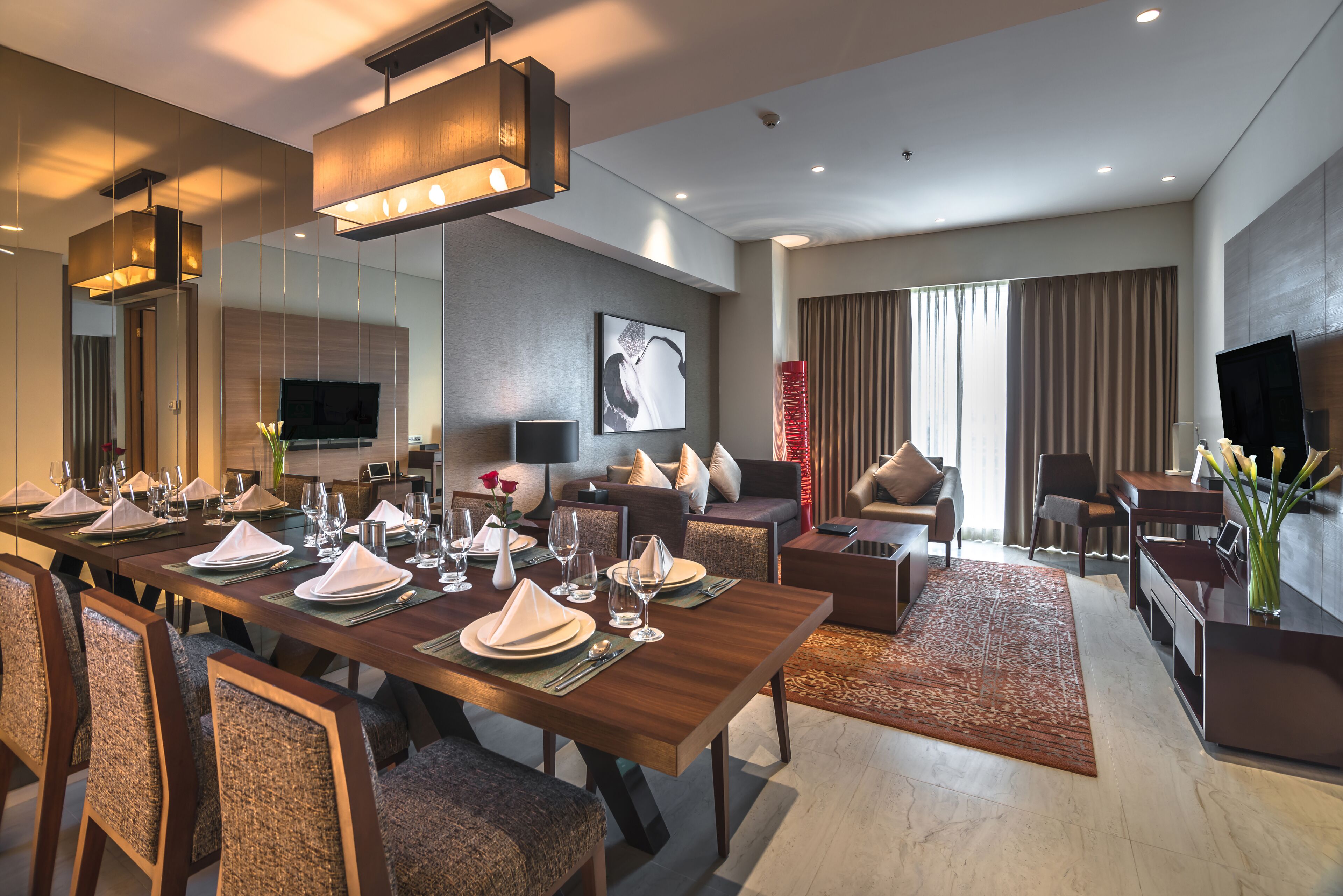 A modern hotel room with a dining area, a living area with a sofa and armchairs, a flat-screen TV, and a large window with curtains.
