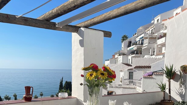 Ein Essbereich im Freien mit Tisch für vier Personen, gelbe Korbstühle, einer Blumenvase und Blick auf das Meer sowie weiße Gebäude.