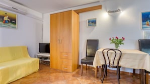 A room with a yellow sofa, a wooden wardrobe, a dining table with chairs, and a TV on a stand.