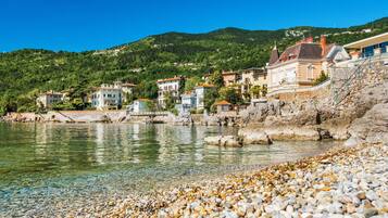 A coastal town with buildings along the shore, a pebbly beach, and a backdrop of green hills.