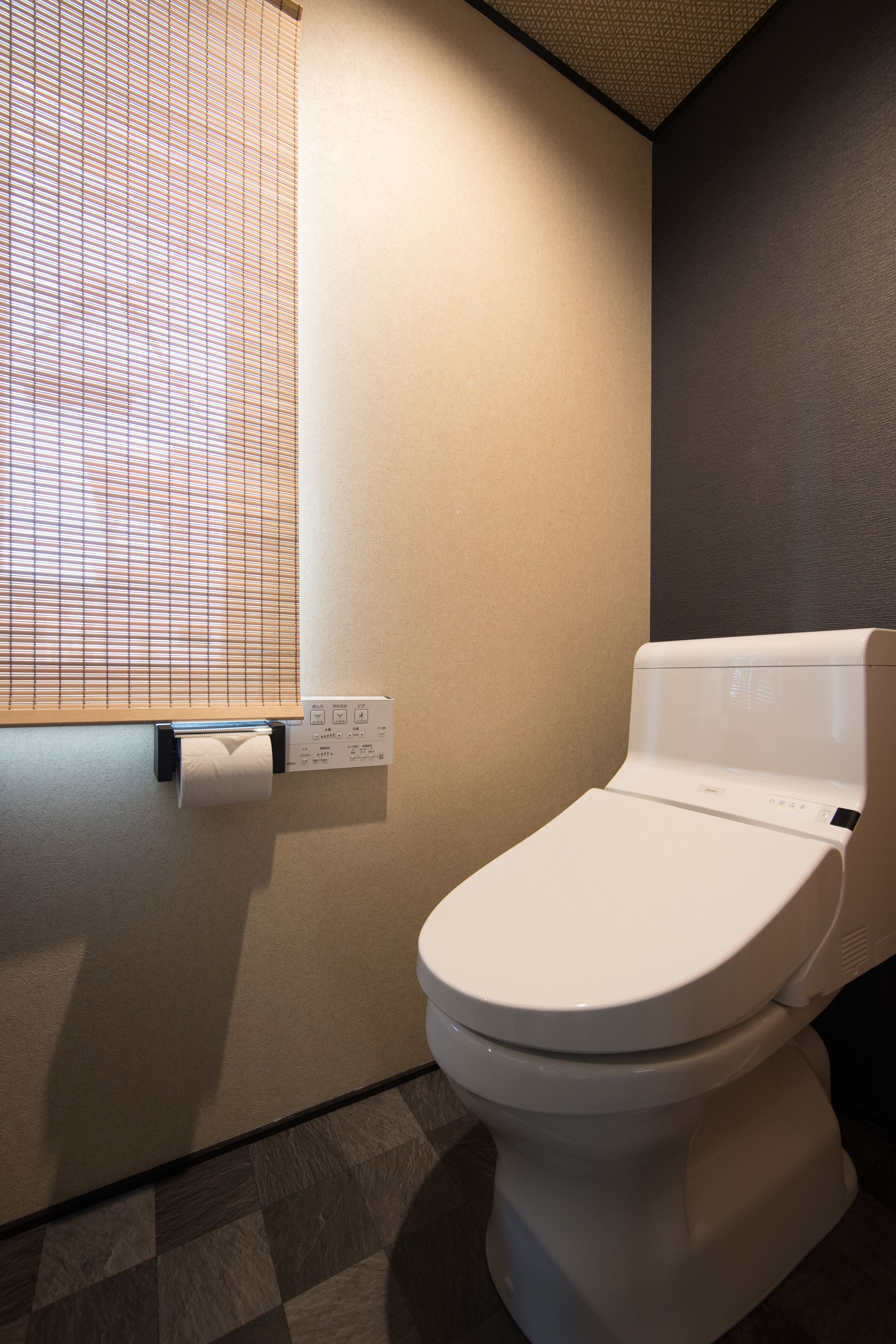 A modern bathroom with a toilet, a paper towel dispenser, and a window with blinds.
