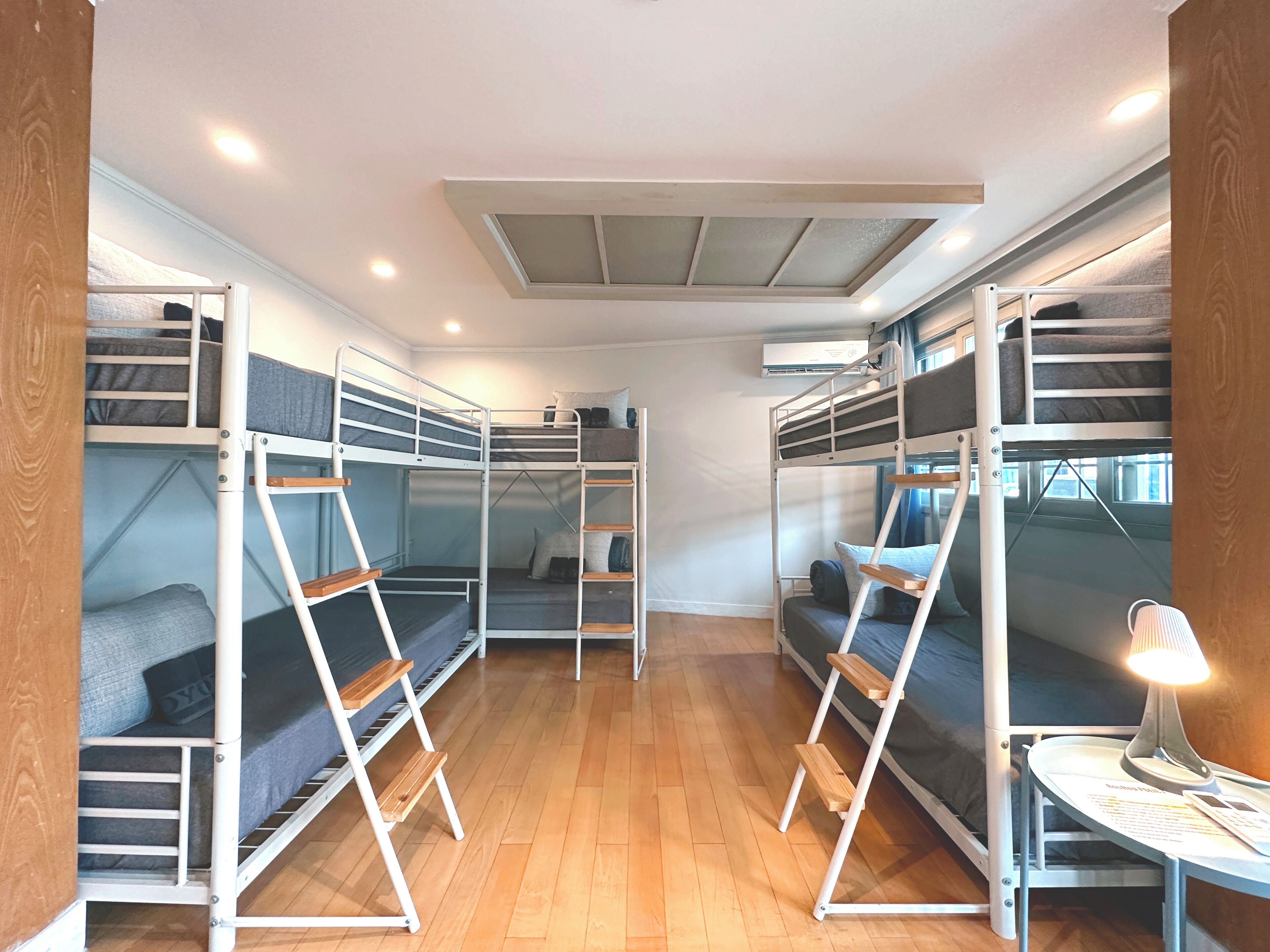 A room with bunk beds, wooden flooring, a skylight, and a table with a lamp.